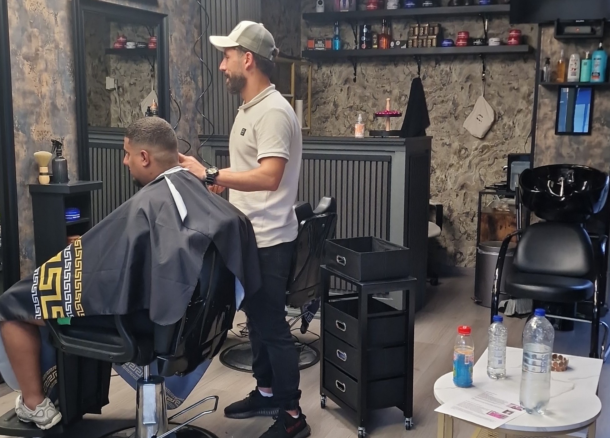 De zaak van binnen: de kapper aan het werk bij een klant in de stoel, met de wasbak en de schappen erachter